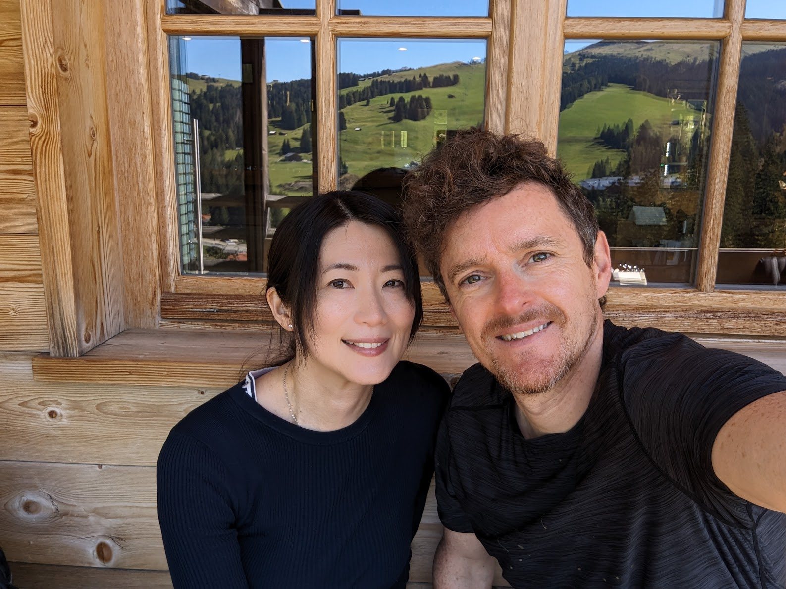 Tom and Rieko Vining outside a wooden chalet in the Dolomites, with green mountain pastures reflected in the window